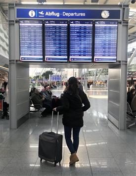 At the Airport. A big blue Screen is listing all departure flights. A Young woman with dark hair, a dark jacket and a small suitcase is standing in front of the screen is looking up.